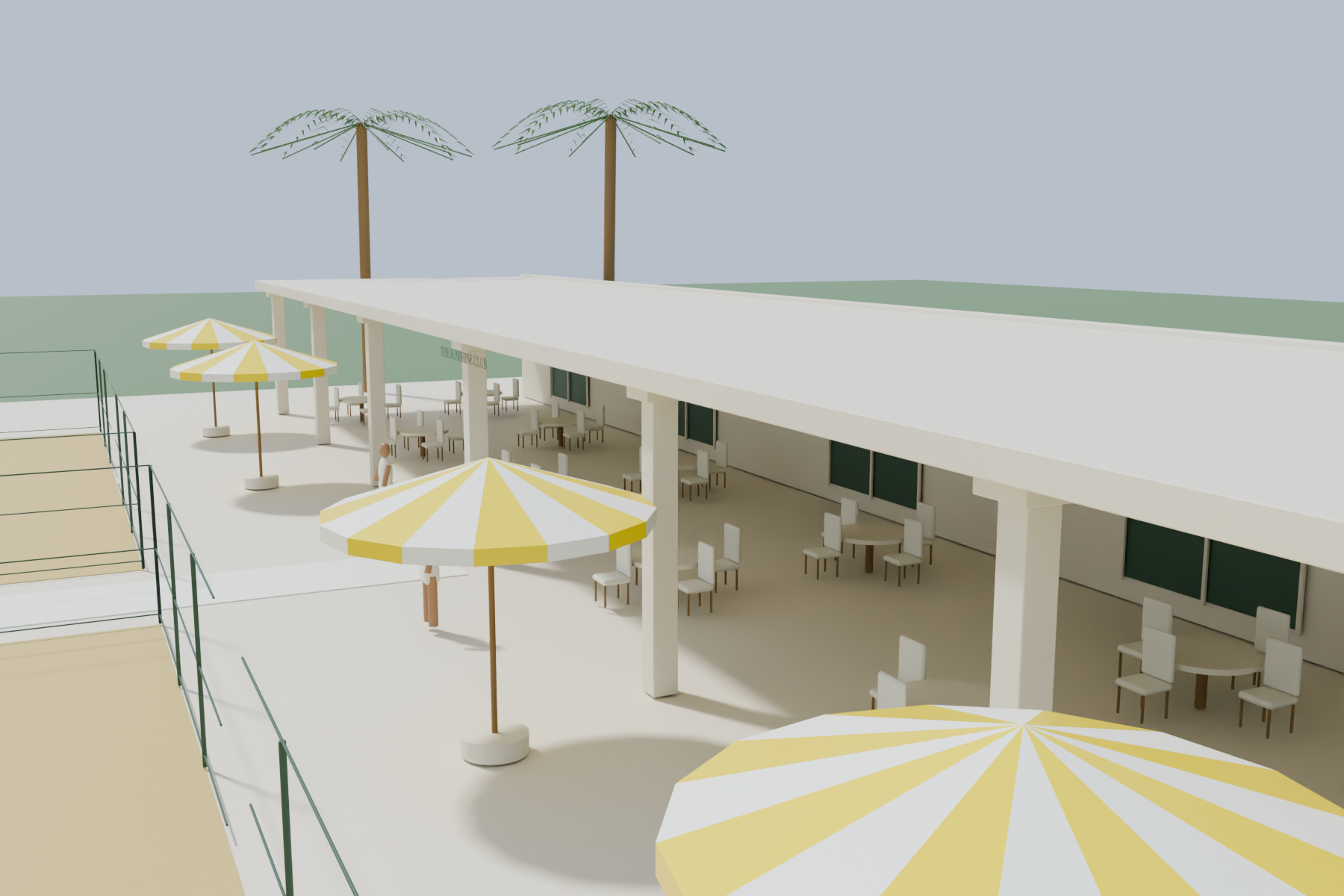 Cream clubhouse veranda with shaded seating, hospitality tables and yellow striped parasols beyond court run-off.